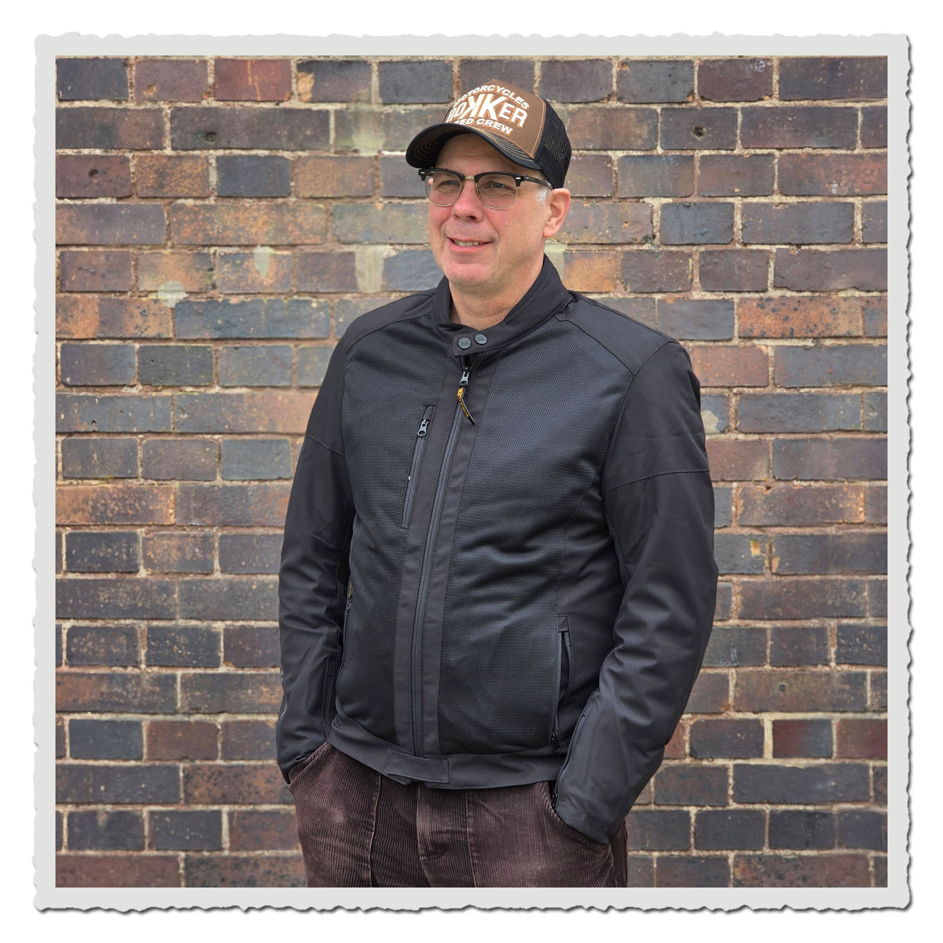 Mann in schwarzer Jacke und brauner Kappe, seitliche Vorderansicht vor brauner Backsteinwand.